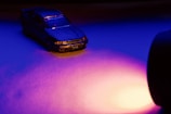 A small toy car is positioned under dramatic lighting, highlighting its details. The scene uses intense blue and purple hues, creating a moody and cinematic atmosphere. A strong light source illuminates from the right, casting dynamic shadows.