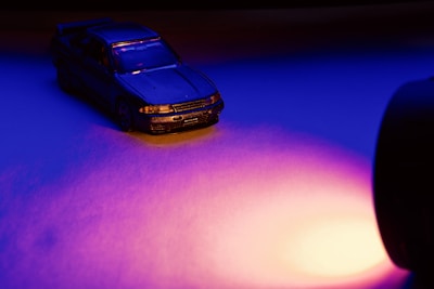 A small toy car is positioned under dramatic lighting, highlighting its details. The scene uses intense blue and purple hues, creating a moody and cinematic atmosphere. A strong light source illuminates from the right, casting dynamic shadows.