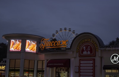 A shopping center at dusk with a large Ferris wheel visible behind it. There are illuminated signs advertising food with images of pastries, and the name of the center displayed in Cyrillic script. The sky is darkening, suggesting evening, and the buildings have various signs with neon lights.