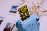 A blue instant camera with a photograph emerging from it. The photograph features a field of yellow flowers. In the blurred background, more photos and some dried plants are visible, creating a soft focus effect.