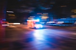 An ambulance with flashing lights speeds down a city street at night. The image captures motion blur, creating a dynamic sense of urgency and movement. The surrounding lights from buildings and street lamps add a blurred effect, emphasizing the speed of the vehicle.