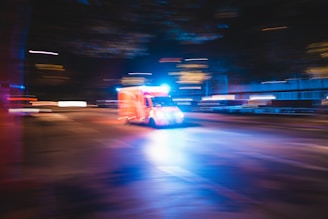An ambulance with flashing lights speeds down a city street at night. The image captures motion blur, creating a dynamic sense of urgency and movement. The surrounding lights from buildings and street lamps add a blurred effect, emphasizing the speed of the vehicle.