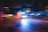 An ambulance with flashing lights speeds down a city street at night. The image captures motion blur, creating a dynamic sense of urgency and movement. The surrounding lights from buildings and street lamps add a blurred effect, emphasizing the speed of the vehicle.