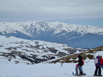 A snowy mountain landscape with a group of people engaged in winter sports. The foreground features several individuals dressed in colorful ski gear, equipped with skis and poles. The background showcases expansive snow-covered mountains under a cloudy sky, creating a serene and vast winter setting.