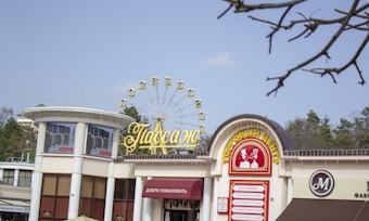 A marketplace or shopping center with a sign in Cyrillic script featuring a golden Ferris wheel and decorative light strings. The building has an outdoor walkway and various shop signs. Trees are visible in the background under a clear sky, and some bare branches are in the foreground.