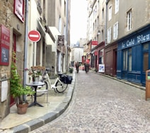 A narrow cobblestone street flanked by historic buildings with shops and cafes. A bicycle is parked near a small table with a flower pot, and potted plants line the walkway. A 'Hotel San Pedro' sign is visible on one building. Farther down the street, a few people are walking near an outdoor cafe.
