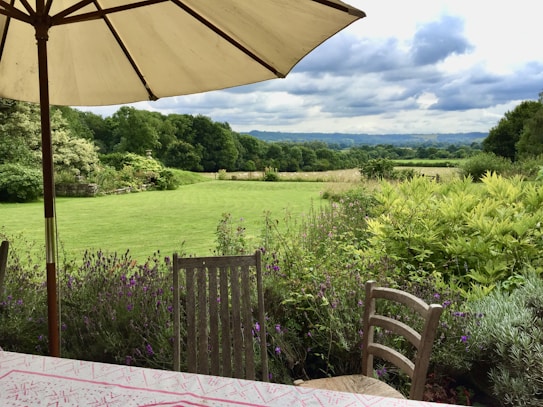 A picturesque outdoor garden scene with a green lawn and surrounding trees in the background. A patio table with a patterned tablecloth and wooden chairs is positioned under a large umbrella. There are vibrant flowers and lush greenery, giving the space a tranquil, inviting feel.