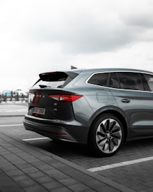 A modern SUV is parked in an outdoor parking lot. The vehicle has a sleek, aerodynamic design with clear emphasis on the sharp lines and large alloy wheels. The rear section is prominently visible, showcasing the distinctive taillights and a partially open trunk. The sky is overcast, adding a moody tone to the setting.