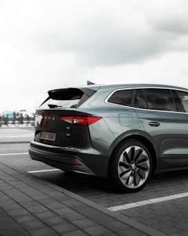 A modern SUV is parked in an outdoor parking lot. The vehicle has a sleek, aerodynamic design with clear emphasis on the sharp lines and large alloy wheels. The rear section is prominently visible, showcasing the distinctive taillights and a partially open trunk. The sky is overcast, adding a moody tone to the setting.