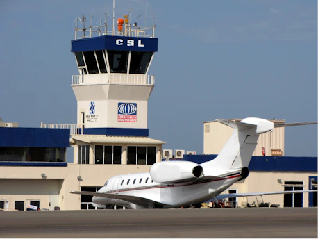 A small airport control tower and terminal building with white and blue colors. A private jet is parked in the foreground on the runway. The control tower has various antennas and equipment on its roof, and the terminal has multiple windows and a logo on display.
