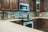A modern kitchen with polished wooden cabinets and stainless steel appliances. The backsplash features neutral-colored stone tiles, and there is a granite countertop. A microwave is mounted above the stove, and a stainless steel kettle sits nearby. A spice rack and a couple of jars are also noticeable.