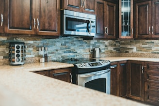 A modern kitchen with polished wooden cabinets and stainless steel appliances. The backsplash features neutral-colored stone tiles, and there is a granite countertop. A microwave is mounted above the stove, and a stainless steel kettle sits nearby. A spice rack and a couple of jars are also noticeable.