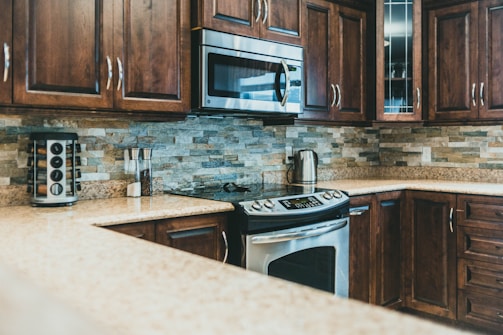 A modern kitchen with polished wooden cabinets and stainless steel appliances. The backsplash features neutral-colored stone tiles, and there is a granite countertop. A microwave is mounted above the stove, and a stainless steel kettle sits nearby. A spice rack and a couple of jars are also noticeable.