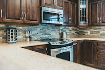 A modern kitchen with polished wooden cabinets and stainless steel appliances. The backsplash features neutral-colored stone tiles, and there is a granite countertop. A microwave is mounted above the stove, and a stainless steel kettle sits nearby. A spice rack and a couple of jars are also noticeable.