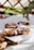 Three freshly baked pastries with a twisted design are topped with sliced almonds and dusted with powdered sugar. They rest on white parchment paper atop a wooden cutting board. Blueberries are scattered around, and there's foliage in the background, giving a fresh and natural ambiance.