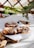 Three freshly baked pastries with a twisted design are topped with sliced almonds and dusted with powdered sugar. They rest on white parchment paper atop a wooden cutting board. Blueberries are scattered around, and there's foliage in the background, giving a fresh and natural ambiance.