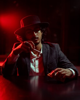 A person wearing a dark hat and glasses sits at a table with a serious expression. The ambient lighting is dim with a strong red hue, creating a moody atmosphere. The individual is holding a pack of cigarettes and a lighter, seemingly about to light one. The attire includes a dark blazer over a white shirt, adding to the sophisticated yet mysterious vibe.