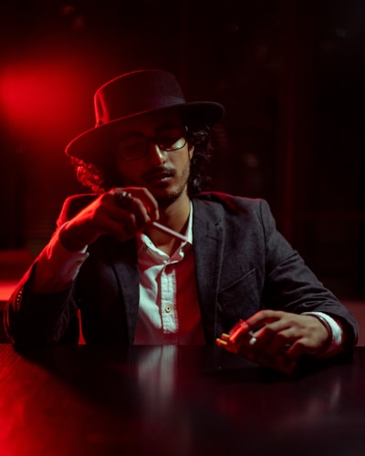 A person wearing a dark hat and glasses sits at a table with a serious expression. The ambient lighting is dim with a strong red hue, creating a moody atmosphere. The individual is holding a pack of cigarettes and a lighter, seemingly about to light one. The attire includes a dark blazer over a white shirt, adding to the sophisticated yet mysterious vibe.