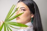 A woman with long dark hair is gazing at the camera while holding a green palm leaf in front of her face. She is wearing makeup with prominent eyelashes, and is adorned with teal earrings and a chunky gold necklace. The background is plain and light-colored.