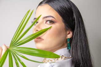 A woman with long dark hair is gazing at the camera while holding a green palm leaf in front of her face. She is wearing makeup with prominent eyelashes, and is adorned with teal earrings and a chunky gold necklace. The background is plain and light-colored.
