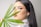 A woman with long dark hair is gazing at the camera while holding a green palm leaf in front of her face. She is wearing makeup with prominent eyelashes, and is adorned with teal earrings and a chunky gold necklace. The background is plain and light-colored.