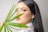 A woman with long dark hair is gazing at the camera while holding a green palm leaf in front of her face. She is wearing makeup with prominent eyelashes, and is adorned with teal earrings and a chunky gold necklace. The background is plain and light-colored.
