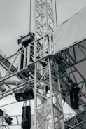 A network of metallic trusses and beams is prominently featured, showcasing an industrial or construction setting. Chains and cables are attached to various parts, with some black equipment hanging from them. A tarpaulin or white fabric covers part of the structure, suggesting an outdoor or temporary installation.