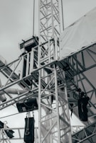 A network of metallic trusses and beams is prominently featured, showcasing an industrial or construction setting. Chains and cables are attached to various parts, with some black equipment hanging from them. A tarpaulin or white fabric covers part of the structure, suggesting an outdoor or temporary installation.