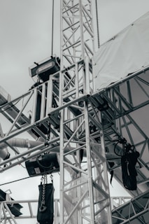 A network of metallic trusses and beams is prominently featured, showcasing an industrial or construction setting. Chains and cables are attached to various parts, with some black equipment hanging from them. A tarpaulin or white fabric covers part of the structure, suggesting an outdoor or temporary installation.