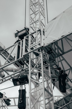 A network of metallic trusses and beams is prominently featured, showcasing an industrial or construction setting. Chains and cables are attached to various parts, with some black equipment hanging from them. A tarpaulin or white fabric covers part of the structure, suggesting an outdoor or temporary installation.
