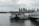 A small structure sits on a platform in a body of water with a metal bridge leading to it. In the background, a city skyline with tall buildings rises under a cloudy sky.