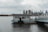 A small structure sits on a platform in a body of water with a metal bridge leading to it. In the background, a city skyline with tall buildings rises under a cloudy sky.