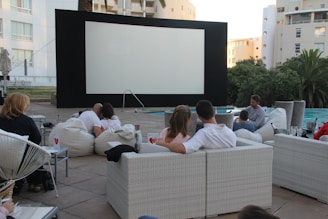 A group of people are sitting on comfortable outdoor seating, including white woven sofas and bean bags, facing an empty large screen set up for a movie screening. The setting is an open-air environment with a swimming pool beside the seating area. The background shows residential buildings and greenery, giving a relaxed, casual vibe.