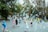 Children are joyfully playing with numerous soap bubbles in a park setting. Surrounding them are large trees with green and yellowing leaves. Adults and other children watch or participate in the fun on a paved pathway.