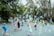 Children are joyfully playing with numerous soap bubbles in a park setting. Surrounding them are large trees with green and yellowing leaves. Adults and other children watch or participate in the fun on a paved pathway.