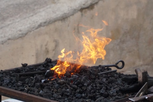 Bright orange flames rise from a bed of black coal or charcoal with metal tools resting nearby, set against a neutral concrete backdrop.