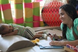 A woman with curly hair is sitting at a table, smiling and writing on a notepad with a marker. Next to her, a young child is peacefully sleeping on a padded surface, wearing a striped shirt and white shorts. In the background, colorful soft blocks and a large red exercise ball with a netting enclosure add to the playful and relaxed setting.