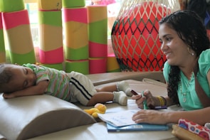A woman with curly hair is sitting at a table, smiling and writing on a notepad with a marker. Next to her, a young child is peacefully sleeping on a padded surface, wearing a striped shirt and white shorts. In the background, colorful soft blocks and a large red exercise ball with a netting enclosure add to the playful and relaxed setting.