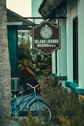 A rustic sign with a decorative compass design hangs outside a building, indicating the location as Island Lounge in the Maldives. Below the sign, a classic blue bicycle is parked amidst lush green foliage, creating a serene and tropical ambiance.