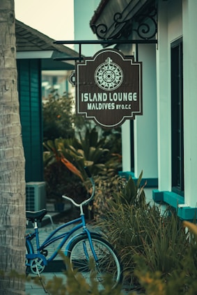A rustic sign with a decorative compass design hangs outside a building, indicating the location as Island Lounge in the Maldives. Below the sign, a classic blue bicycle is parked amidst lush green foliage, creating a serene and tropical ambiance.