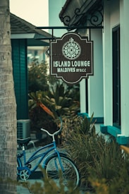 A rustic sign with a decorative compass design hangs outside a building, indicating the location as Island Lounge in the Maldives. Below the sign, a classic blue bicycle is parked amidst lush green foliage, creating a serene and tropical ambiance.