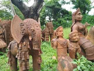 Terracotta sculptures featuring intricately detailed figures are displayed outdoors among lush greenery. The sculptures include an elephant with ornate carvings, alongside humanoid figures adorned with elaborate jewelry and headgear. These artworks are crafted with a rich, earthy brown tone.