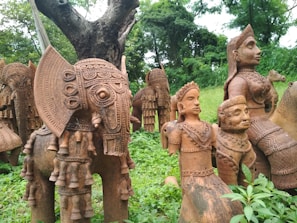 Terracotta sculptures featuring intricately detailed figures are displayed outdoors among lush greenery. The sculptures include an elephant with ornate carvings, alongside humanoid figures adorned with elaborate jewelry and headgear. These artworks are crafted with a rich, earthy brown tone.