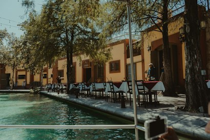 A calm outdoor restaurant setting beside a water body, with multiple tables and chairs arranged under the shade of large trees. Two people are visible near the restaurant entrance, which is styled with warm yellow and brown colors. Sunlight filters through the trees, creating dappled shadows on the ground.