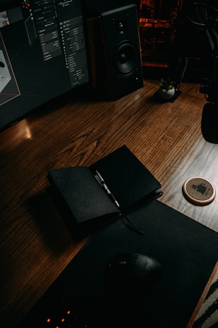 A dark, moody workspace features a wooden desk with an open black notebook and pen. A computer monitor displaying a design program is visible, alongside a large speaker. A potted plant and a drink coaster are also on the desk, with ambient red lighting in the background.