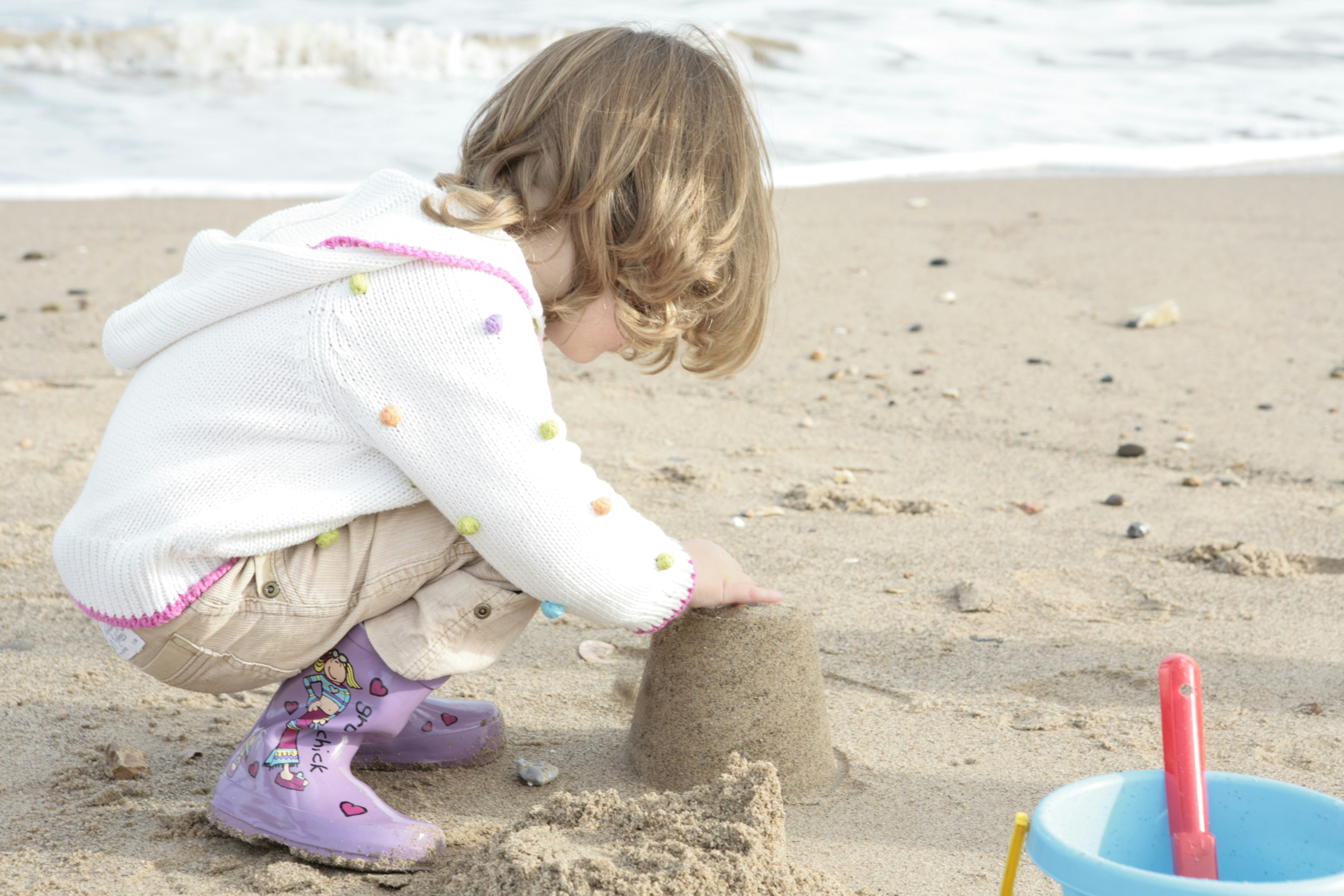 girl in white and pink polka dot jacket sitting on brown sand during daytime, Little girl making a sandcastle at seaside