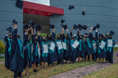 A group of graduates wearing black robes and teal sashes throw their caps in the air while holding certificates. They are standing on a grassy area in front of a modern building with grey and red elements.