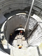 Three workers wearing orange uniforms and yellow helmets are inside a large, cylindrical concrete structure, possibly a construction site or a well. They are maneuvering a wooden platform or scaffold. Sunlight casts long shadows inside the structure, illuminating the damp, grey walls and muddy floor.