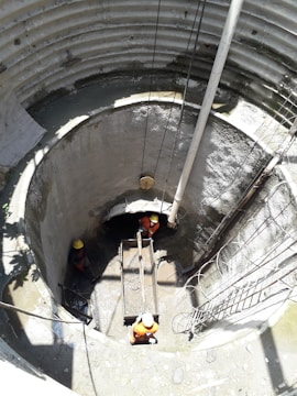 Three workers wearing orange uniforms and yellow helmets are inside a large, cylindrical concrete structure, possibly a construction site or a well. They are maneuvering a wooden platform or scaffold. Sunlight casts long shadows inside the structure, illuminating the damp, grey walls and muddy floor.