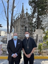 Two men wearing face masks stand in front of an ornate, Gothic-style mausoleum surrounded by trees. One man is wearing a plaid suit jacket and the other is dressed in clergy attire with a cross necklace. A sign in Spanish is visible on the right side.